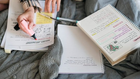 Student studies for exam, with numerous notebooks on her bed