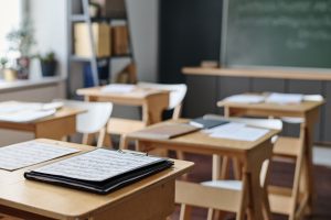 An empty classroom with wooden desks, notebooks on clipboards, chairs, and a chalkboard in the background.